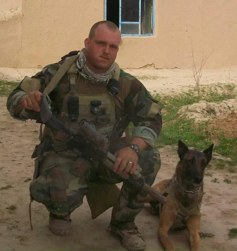 Jacob Robinson in Marine Corps uniform crouching beside a Belgian Malinois in a desert environment